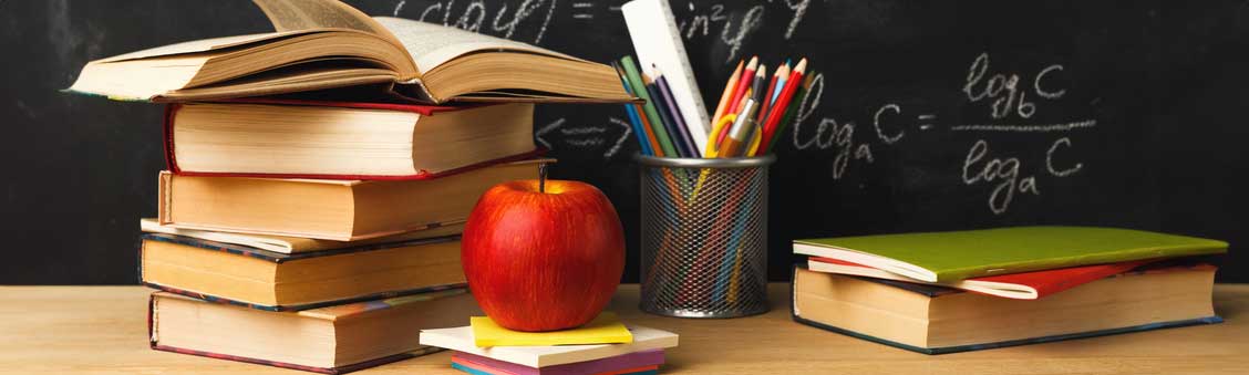 A stack of school books on a desk in front of a chalkboard next to an apple.