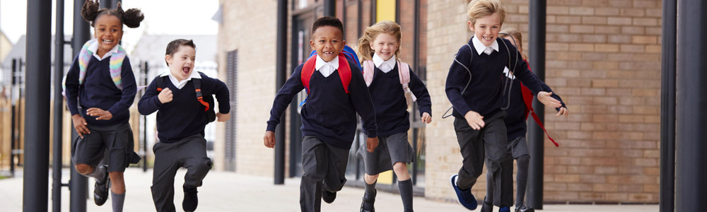 Primary school kids, wearing school uniforms and backpacks, running on a walkway outside their school building, front view