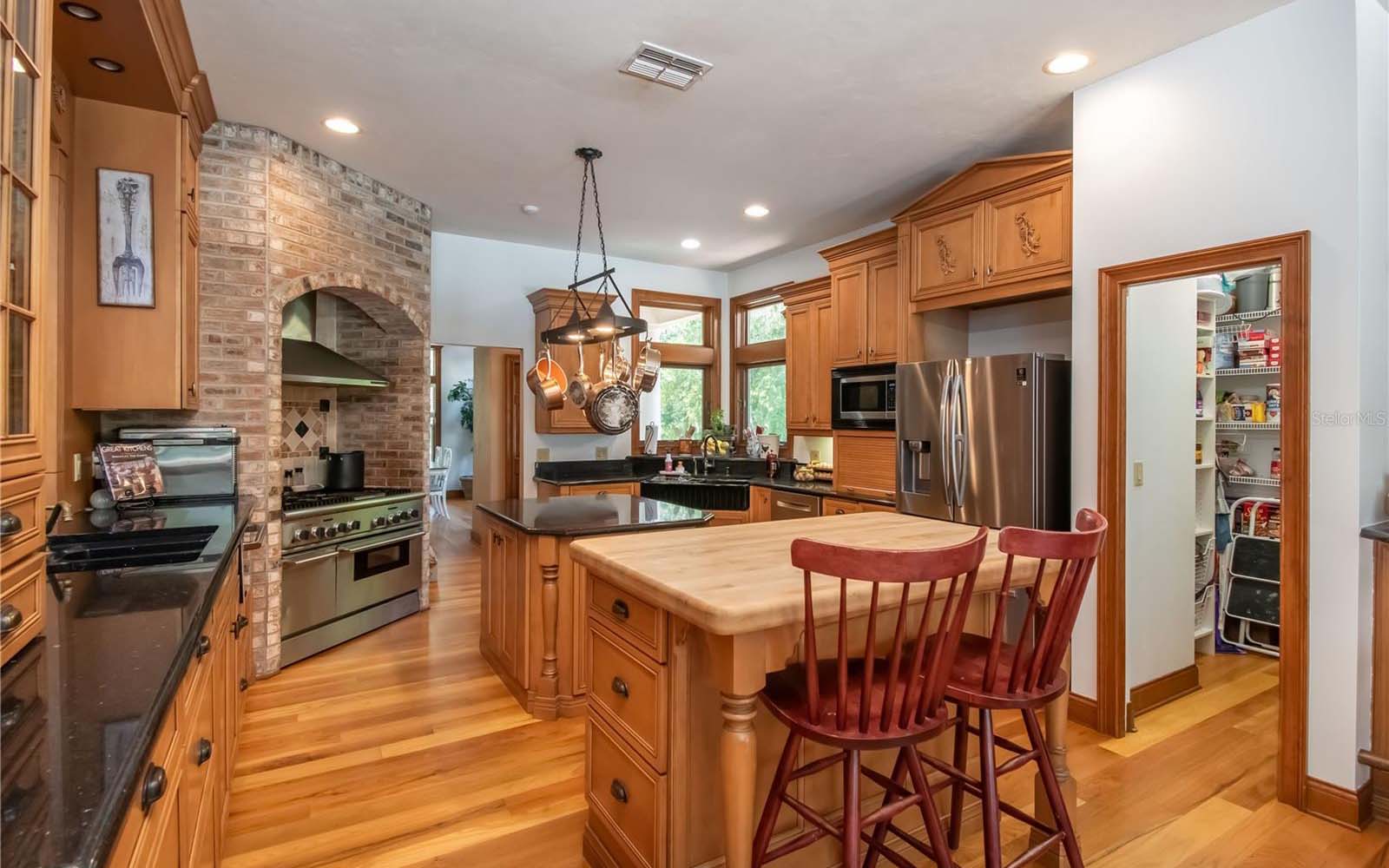 A beautiful kitchen with wood cabinetry, a brick oven alcove, and ornate pot rack.